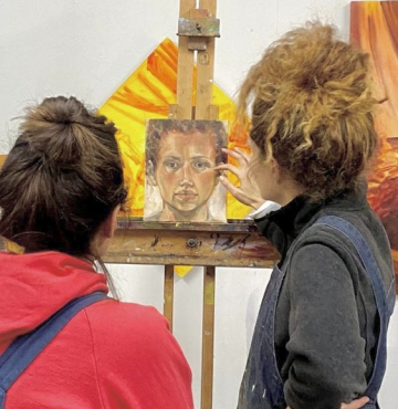 Two women discussing a small portrait painting on an easel.