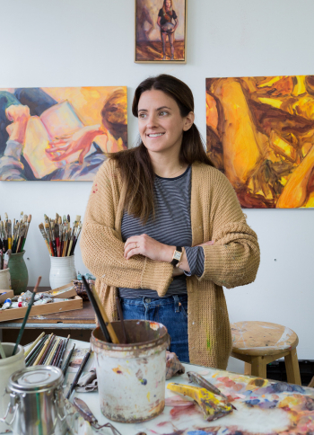 Lee in her studio, smiling, with paintbrushes and artworks on the wall.